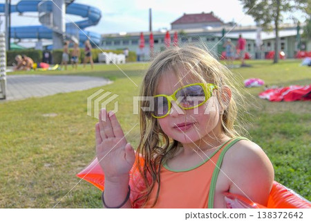 Young girl with sunglasses waving hello at a water park on a sunny day. A cheerful young girl wearing bright sunglasses and an orange swimsuit waves her hand while sitting on inflatable armbands at a Young girl with sunglasses waving hello at a water park on a sunny day. A cheerful young girl wearing bright sunglasses and an orange swimsuit waves her hand while sitting on inflatable armbands at a 138372642