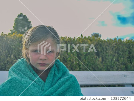 A sad little girl covered in a towel sits on a bench at the swimming pool. The concept of summer vacations, freedom and childhood. A sad little girl covered in a towel sits on a bench at the swimming pool. The concept of summer vacations, freedom and childhood. 138372644