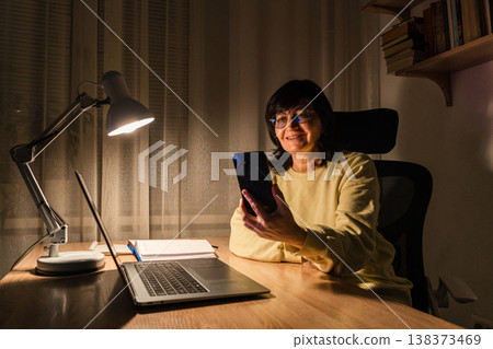 Mature woman in glasses working from home night, smiling while using smartphone laptop at desk with lamp light, working remotely Mature woman in glasses working from home night, smiling while using smartphone laptop at desk with lamp light, working remotely 138373469