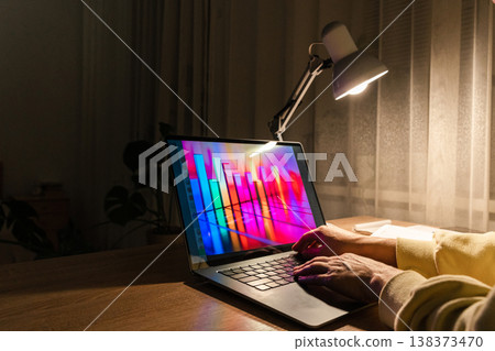 Hands typing on laptop keyboard with colorful bar chart displaying on screen, illuminated by desk lamp at night. Person working at night with laptop showing data chart Hands typing on laptop keyboard with colorful bar chart displaying on screen, illuminated by desk lamp at night. Person working at night with laptop showing data chart 138373470