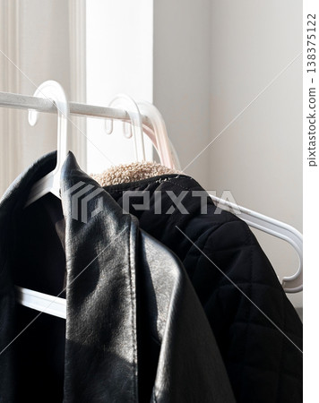 Two jackets hang from white plastic hangers. Smooth texture and quilted design. Hangers placed against light wall, bright sunlight comes through nearby window. Organization and interior 138375122