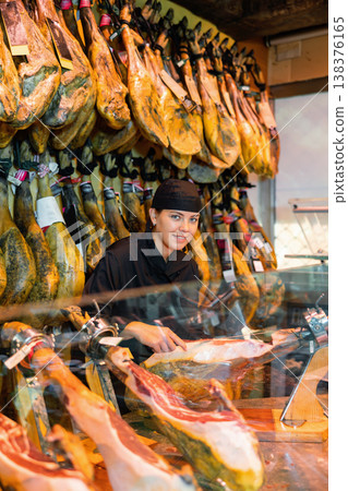 Young female seller in black uniform slicing jamon on jamonera in butcher shop Young female seller in black uniform slicing jamon on jamonera in butcher shop 138376165