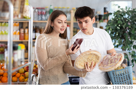 Happy pretty young wife and husband choose large and fresh pizza, scans QR code from label in supermarket in department of semi-finished products Happy pretty young wife and husband choose large and fresh pizza, scans QR code from label in supermarket in department of semi-finished products 138376189