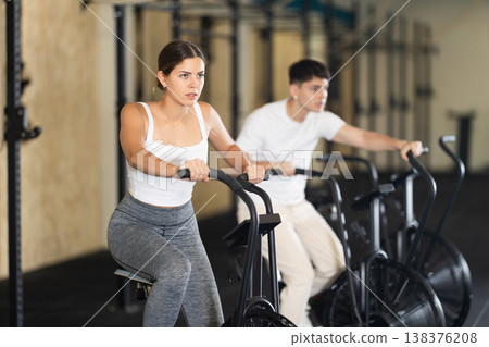 Positive girl riding exercise airbike for cardio workout during cycling class at modern crossfit gym Positive girl riding exercise airbike for cardio workout during cycling class at modern crossfit gym 138376208