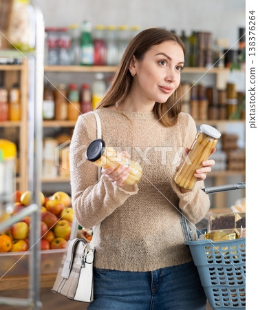Young woman customer with shopping basket buying food in grocery store, reading ingredients on jar of conserve asparagus Young woman customer with shopping basket buying food in grocery store, reading ingredients on jar of conserve asparagus 138376264