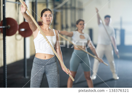 Slender girl, together with other athletes, does a warm-up using a long stick in gym Slender girl, together with other athletes, does a warm-up using a long stick in gym 138376321