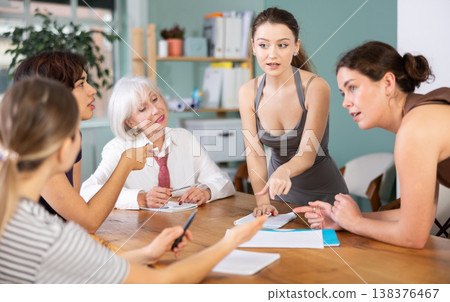 Positive young girl office worker standing with pen in hands near table where sitting colleagues with papers and talking in office 138376467