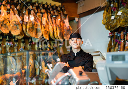 Young butcher shop salesman in black uniform standing with crossed arms near rack with hanging jamon legs Young butcher shop salesman in black uniform standing with crossed arms near rack with hanging jamon legs 138376468