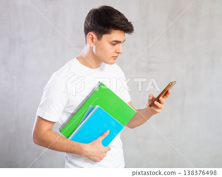 Attentive young student with workbooks in hands looking at mobile phone Attentive young student with workbooks in hands looking at mobile phone 138376498