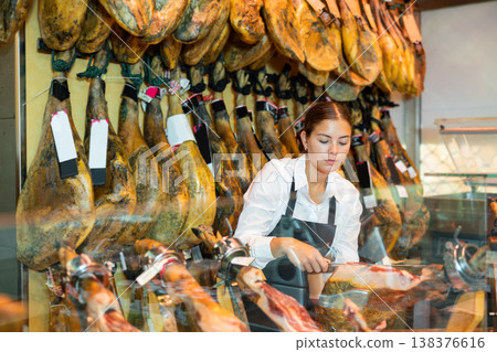 Young saleswoman in black apron slicing Iberian jamon at counter in butcher shop Young saleswoman in black apron slicing Iberian jamon at counter in butcher shop 138376616