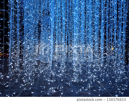 Stunning curtain of blue LED string lights hanging vertically in a dark setting. Festive holiday decoration or immersive light installation with bright glowing bulbs creating a magical atmosphere. Stunning curtain of blue LED string lights hanging vertically in a dark setting. Festive holiday decoration or immersive light installation with bright glowing bulbs creating a magical atmosphere. 138376833