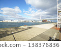 Wide view of a concrete harbor pier and waterfront featuring a large cable-stayed bridge under a cloudy blue sky. Industrial buildings and a cargo ship line the bay in Osaka, Japan. Urban scenery. 138376836