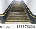 Symmetrical low-angle view of stone stairs leading upward in an underground subway station. Bright light glows at the top. Features unique yellow wavy handrails against dark and white walls. 138376839