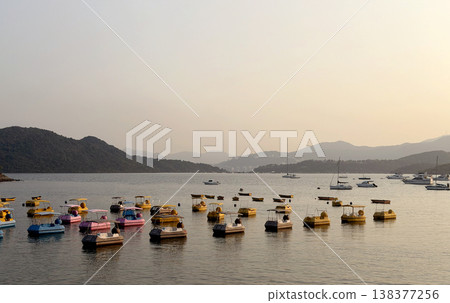 Colorful paddle boats and sailboats dot a calm bay at sunset with rolling hills 138377256