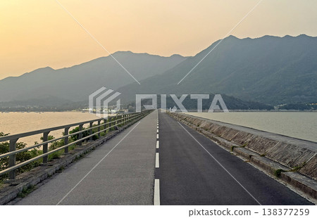 Scenic dam road stretching towards misty mountains at sunset with calm water on both sides Scenic dam road stretching towards misty mountains at sunset with calm water on both sides 138377259