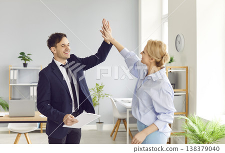 Two joyful colleagues give triumphant high five in office symbolizing achievement and team spirit. Two joyful colleagues give triumphant high five in office symbolizing achievement and team spirit. 138379040