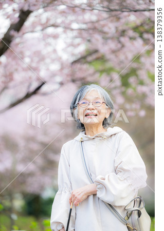 An elderly woman smiling under a cherry tree amidst falling petals evokes an image of health and happiness in spring; a senior woman gazing up at cherry blossoms during a cherry blossom viewing. 138379356
