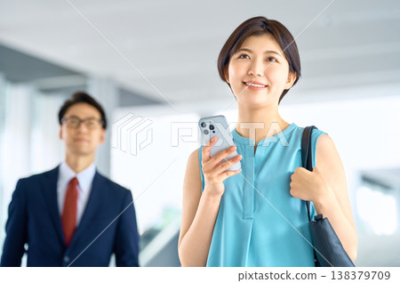 一位面帶笑容的女商人手持智慧型手機，背景中可以看到一位商人。 138379709