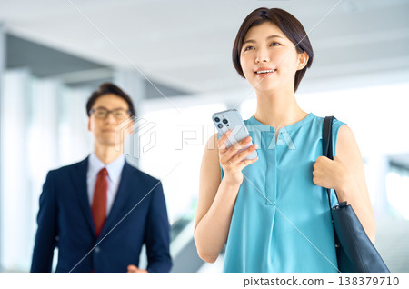 一位微笑的日本女商人與一位身穿西裝的男士走過一棟辦公大樓，兩人都拿著一支智慧型手機。 138379710