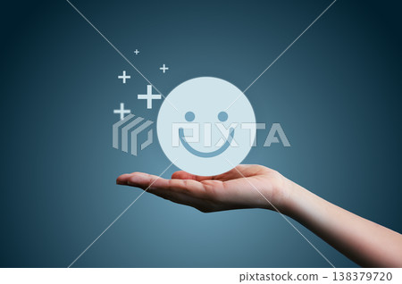 Hand holding smiling face icon with plus signs on dark blue background. Concept of customer satisfaction, positive rating, excellent service feedback, mental health wellbeing, and pure happiness. 138379720