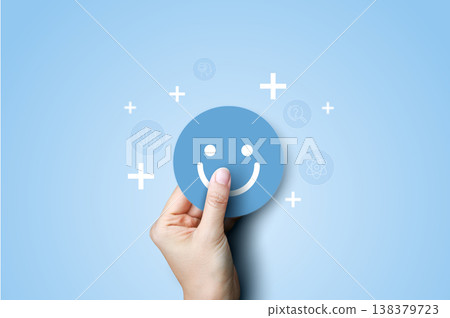 Hand holding smiling face icon with plus signs on blue background. Concept of customer satisfaction, positive feedback, added value, excellent service rating, and mental health wellness. 138379723