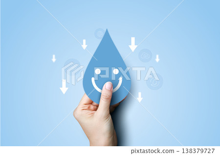 Hand holding a smiling blue water drop icon with downward arrows on blue background. Concept of reducing water consumption, saving water, environmental conservation, and eco-friendly sustainability. 138379727