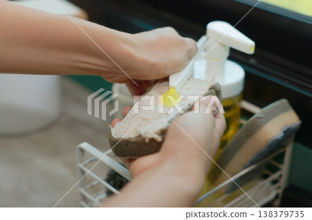 Close up of hands pumping yellow dishwashing liquid soap from a dispenser bottle onto a cleaning sponge at the kitchen sink. Washing dishes, household chores, hygiene and housework daily routine. Close up of hands pumping yellow dishwashing liquid soap from a dispenser bottle onto a cleaning sponge at the kitchen sink. Washing dishes, household chores, hygiene and housework daily routine. 138379735