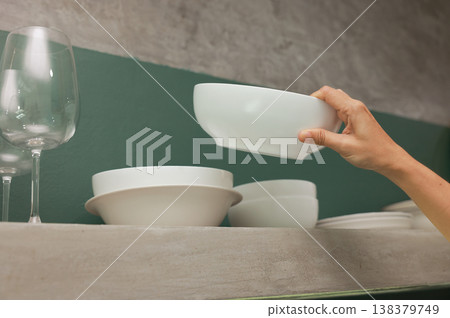 Close up of a person hand organizing clean white ceramic bowls on a modern concrete kitchen shelf against a green wall. Domestic household chores, tidying up and daily home daily routine. Close up of a person hand organizing clean white ceramic bowls on a modern concrete kitchen shelf against a green wall. Domestic household chores, tidying up and daily home daily routine. 138379749