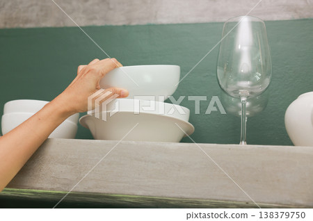 Close up of a person hand organizing clean white ceramic bowls on a modern concrete kitchen shelf against a green wall. Domestic household chores, tidying up and daily home daily routine. Close up of a person hand organizing clean white ceramic bowls on a modern concrete kitchen shelf against a green wall. Domestic household chores, tidying up and daily home daily routine. 138379750
