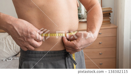 Man measuring his waist with a tape measure, checking progress on his weight loss journey in the bedroom. Real person, self-acceptance, authentic lifestyle Man measuring his waist with a tape measure, checking progress on his weight loss journey in the bedroom. Real person, self-acceptance, authentic lifestyle 138382115