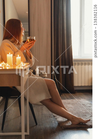 Woman in bathrobe enjoying hot tea illuminated by flickering candles, finding peace and tranquility. Wellness and spa evening Woman in bathrobe enjoying hot tea illuminated by flickering candles, finding peace and tranquility. Wellness and spa evening 138382116