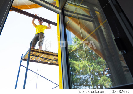 Window reflection shows worker balancing on scaffold, fixing wood trim at height, focus on manual labor, modern house renovation. Window reflection shows worker balancing on scaffold, fixing wood trim at height, focus on manual labor, modern house renovation. 138382652