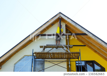 Straight-on view of yellow modern home, scaffold and tools for wooden gable decor, ongoing DIY renovation, exterior structure evident. Straight-on view of yellow modern home, scaffold and tools for wooden gable decor, ongoing DIY renovation, exterior structure evident. 138382663
