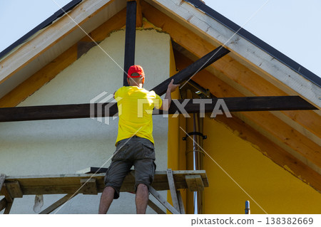 Craftsman reaches high to mount wooden crosspiece under peaked roof, yellow home facade, visible tools, advanced roofing skill. Craftsman reaches high to mount wooden crosspiece under peaked roof, yellow home facade, visible tools, advanced roofing skill. 138382669