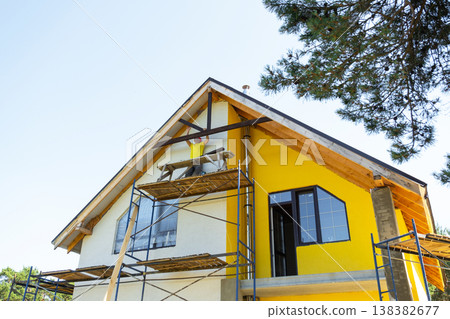 Man works on side of house installing decorative wooden design under roof slope, yellow gable and large window, DIY renovation on height. 138382677