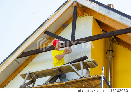 Carpenter adjusts horizontal wooden trim under roof peak, exterior works at height, safety harness, gable decor in progress, yellow residential facade. Carpenter adjusts horizontal wooden trim under roof peak, exterior works at height, safety harness, gable decor in progress, yellow residential facade. 138382680