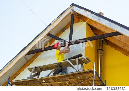 Worker inspects alignment of decorative beams on house gable, hanging from scaffold, modern yellow home, craftsmanship and attention to detail. Worker inspects alignment of decorative beams on house gable, hanging from scaffold, modern yellow home, craftsmanship and attention to detail. 138382681