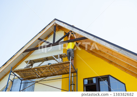 Craftsman in yellow shirt fits custom woodwork to gable peak, minimalist facade, working with measuring tape and toolbelt, DIY architecture focus. 138382682