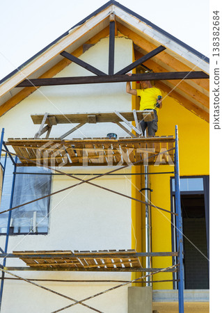 Man in helmet finalizes exterior painting of gable trim, high scaffold, modern yellow house, safety awareness, detail-oriented construction. Man in helmet finalizes exterior painting of gable trim, high scaffold, modern yellow house, safety awareness, detail-oriented construction. 138382684