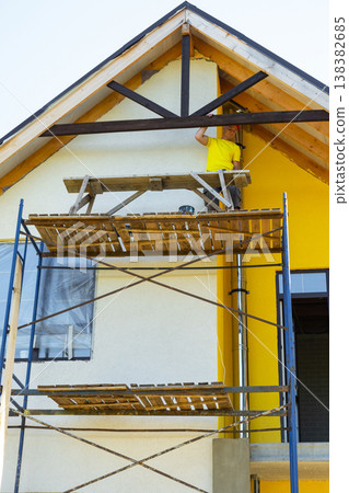 Builder inspects completed wooden gable design, stands on external scaffold, modern yellow facade, craftsmanship achievement, risk on height. Builder inspects completed wooden gable design, stands on external scaffold, modern yellow facade, craftsmanship achievement, risk on height. 138382685