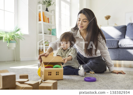 Mother and son playing wooden dollhouse game together at home, sitting on carpet in living room 138382934
