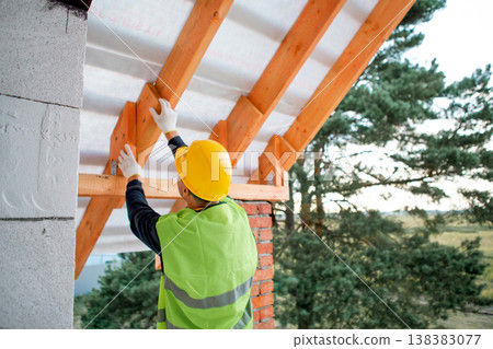 Builder secures roof joist using tool, house under construction with visible timber rafters, assembling main roof framework. Builder secures roof joist using tool, house under construction with visible timber rafters, assembling main roof framework. 138383077