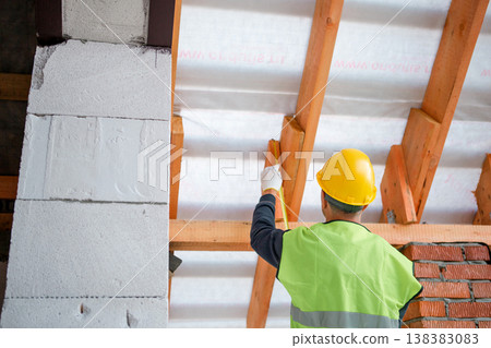 Worker using level tool adjusts wooden rafter during roofing, brick wall backdrop, house structure at roof installation stage. 138383083
