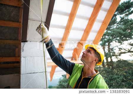 Builder in yellow helmet inspects and prepares uppermost rafters for secure bolting, timber lag connections visible, sunny weather, roof structure. 138383091