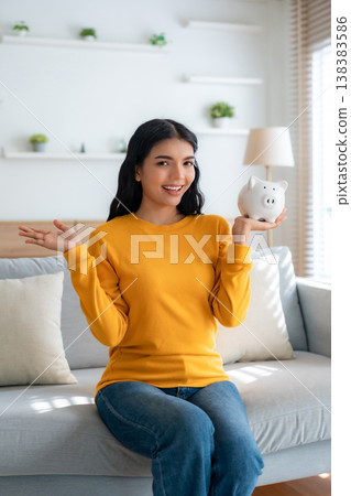 Young Asian woman holding piggy bank and sitting on couch 138383586