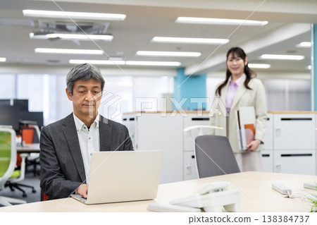 一名男子正在使用筆記型電腦工作，背景中一名女子正在尋找一間空置辦公室的座位。 138384737