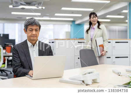 A man is working on his laptop, with a woman in the background looking for a seat in a free-address office. A man is working on his laptop, with a woman in the background looking for a seat in a free-address office. 138384738