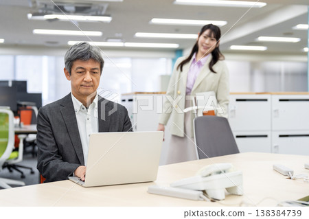 A man is working on his laptop, with a woman in the background looking for a seat in a free-address office. A man is working on his laptop, with a woman in the background looking for a seat in a free-address office. 138384739