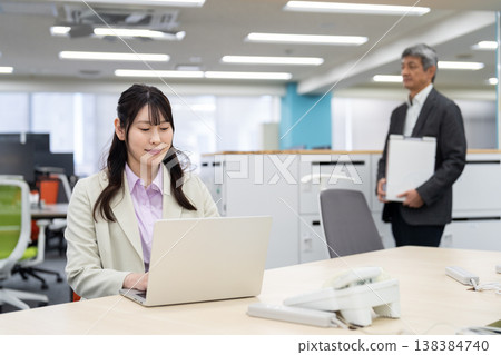 一位女士正在使用筆記型電腦工作,背景中一位男士正在尋找一間空置辦公室的座位。 一位女士正在使用筆記型電腦工作,背景中一位男士正在尋找一間空置辦公室的座位。 138384740