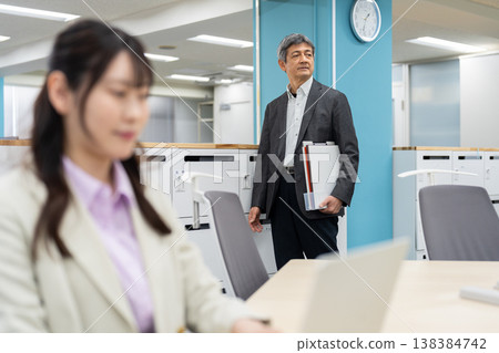 A woman is working on her laptop, with a man in the background looking for a seat in a free-address office. 138384742